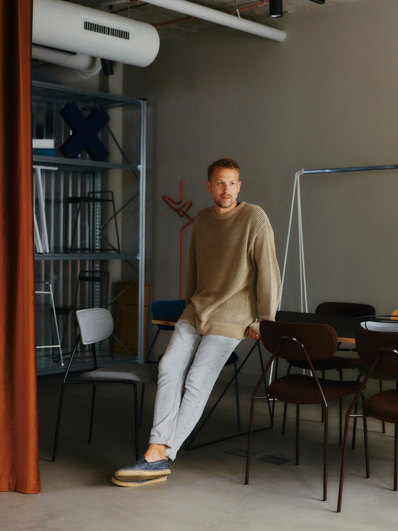 Man in beige sweater sitting casually on black chair in modern office space with glass shelving and ventilation