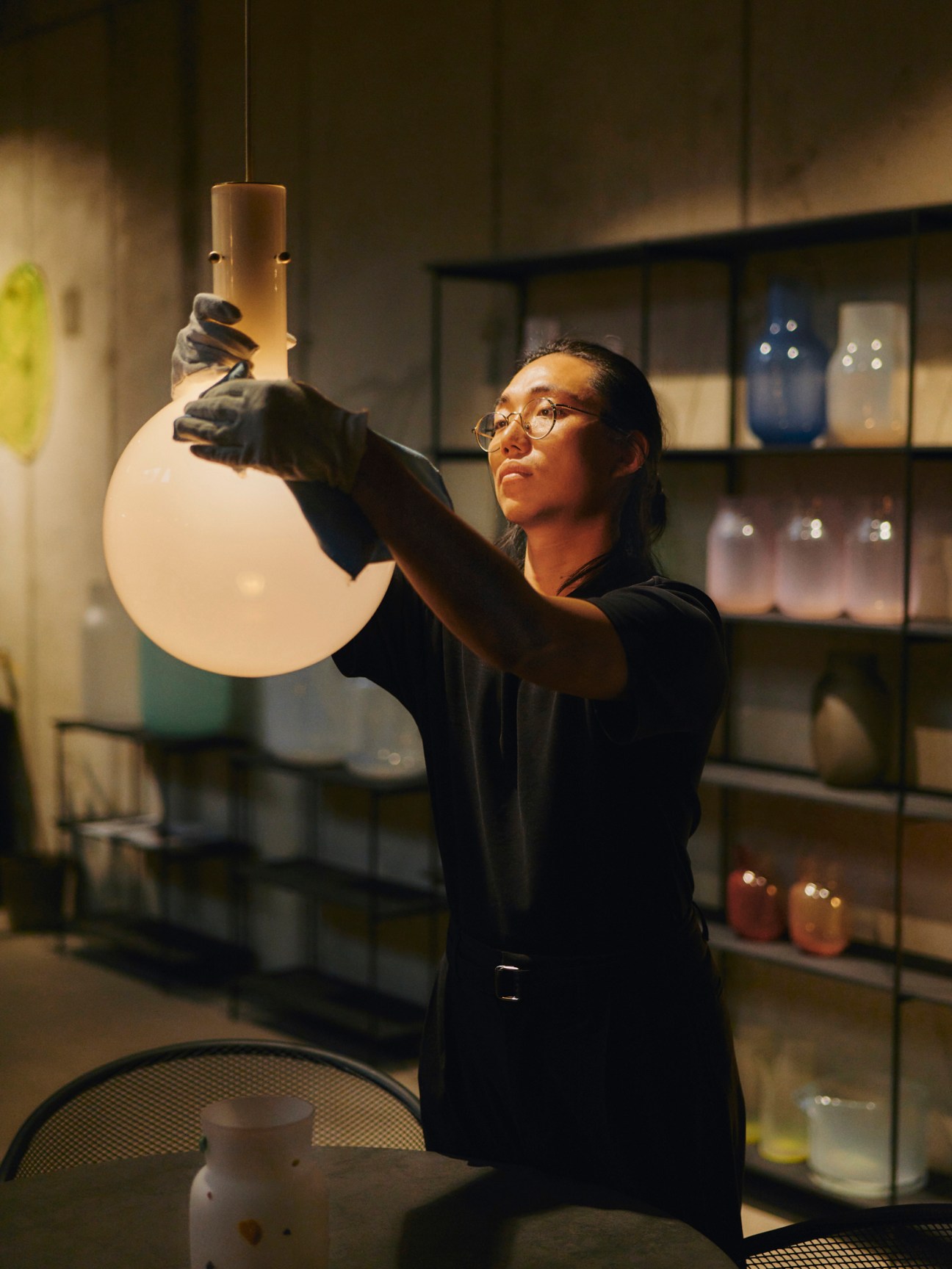 A craftsperson in black clothing and glasses examines a glass vessel while working in a well-equipped studio.