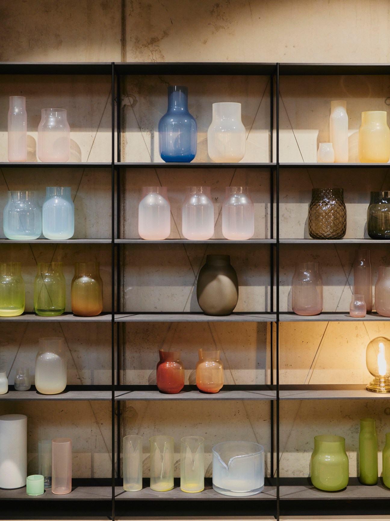Illuminated glass vessels and bottles displayed on black metal shelving against a weathered wall.
