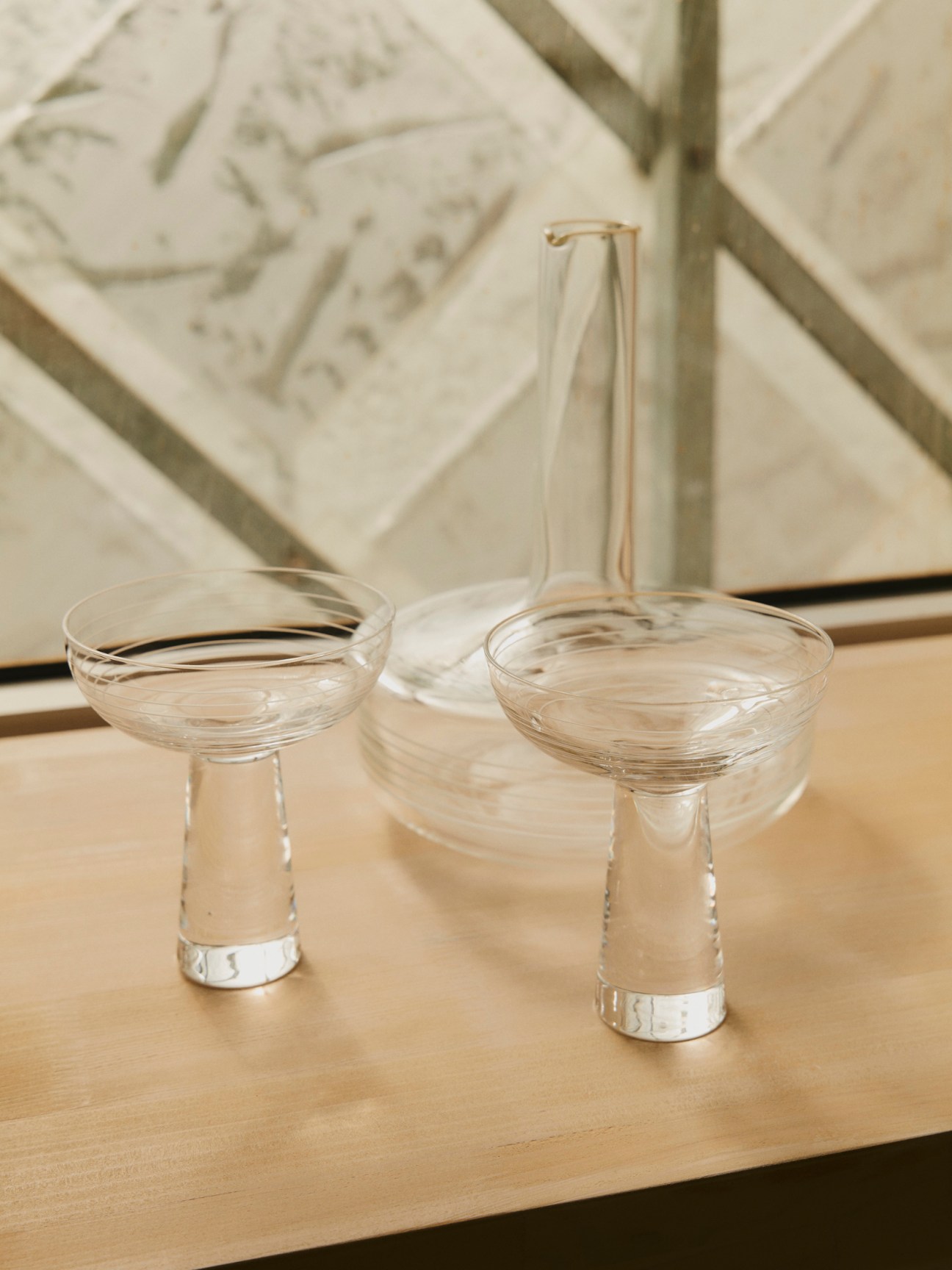 Two clear glass coupe glasses and a glass decanter arranged on a wooden surface with carved reliefs in background