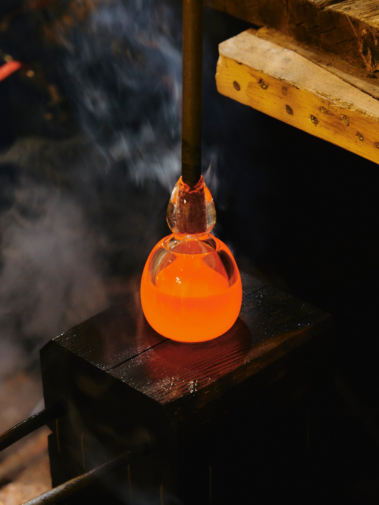 Glassblower at Lasvit creates a glowing orange molten glass form with a tool against a dark background.