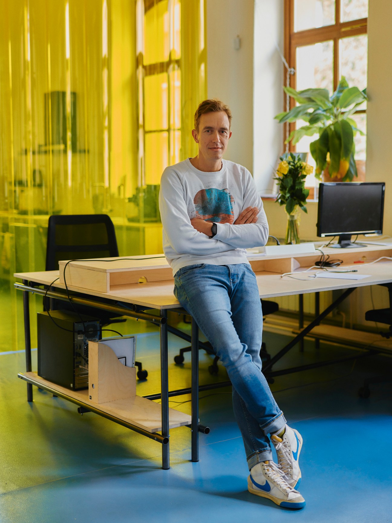 Jan Vondrák sits on a wooden desk in a bright office with yellow glass partitions and modern furniture