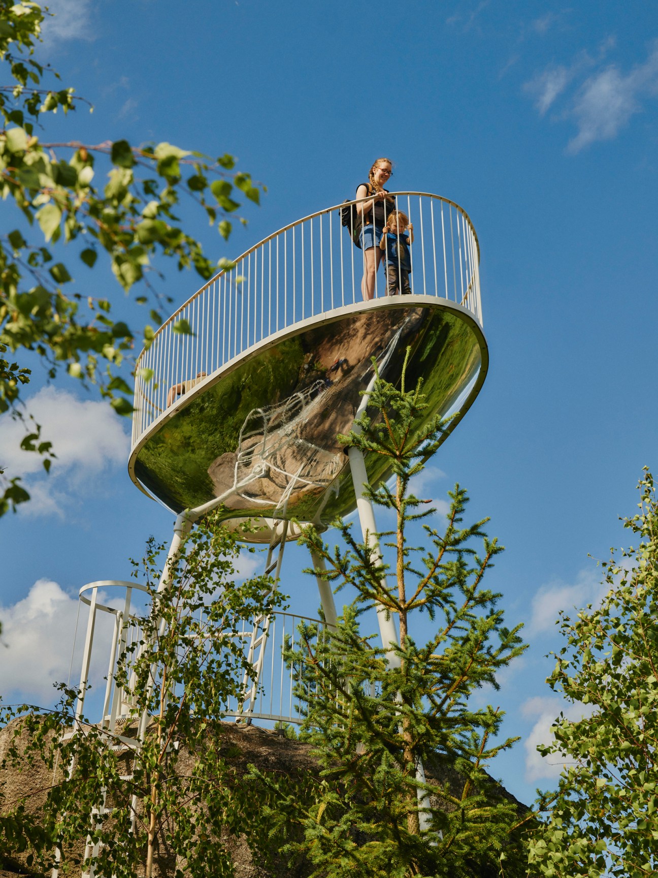 A person stands on a curved metal lookout platform with a reflective underside against a blue sky, surrounded by green vegetation.