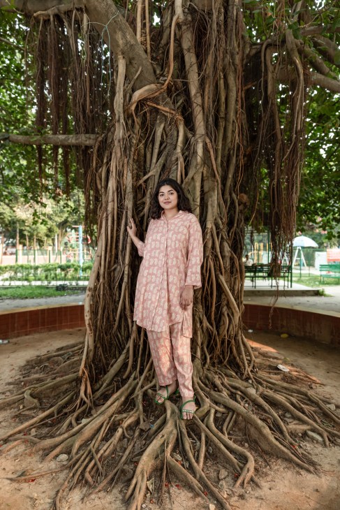 Bushra Afreen stands against large tree roots in pink patterned outfit in urban park setting