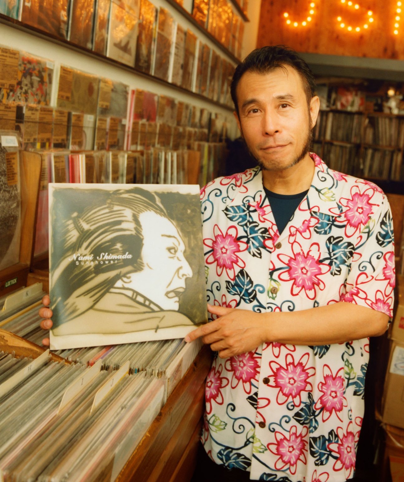 Soichi Terada holds a vinyl record in a record shop surrounded by shelves of music albums.
