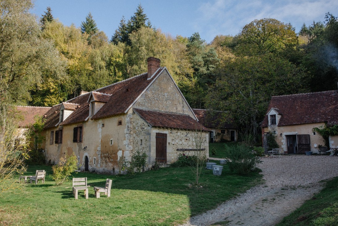 Stone farmhouse with rustic terrace and wooden chairs surrounded by autumn trees at D'une île guesthouse in France.