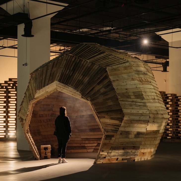 Person standing inside large wooden sculptural installation in warehouse gallery space
