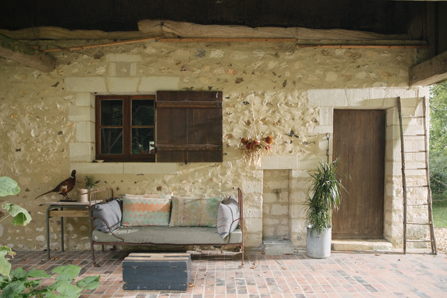 Stone cottage exterior with cushioned seating area, wooden door, and potted plants under shade.