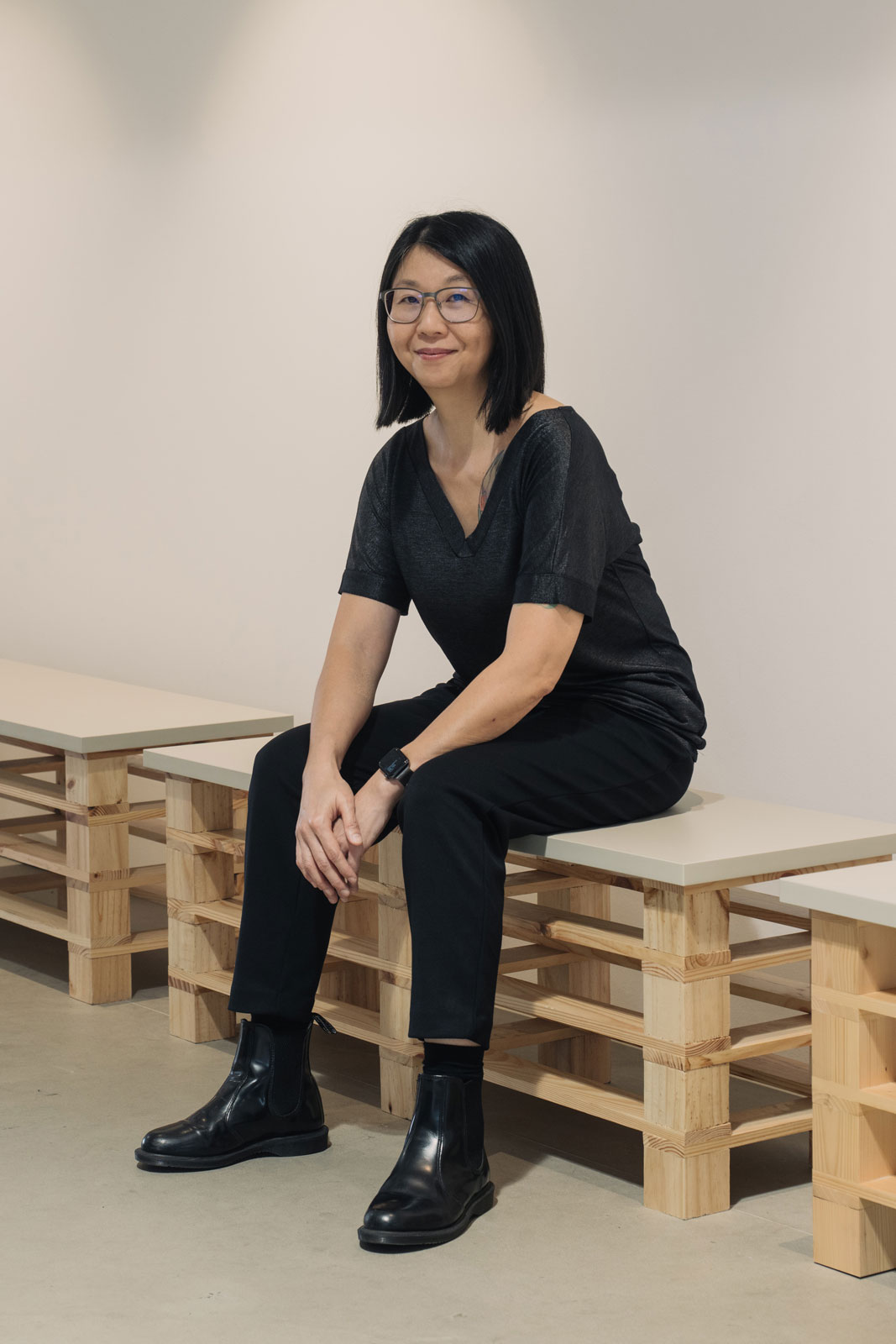 June Yap sitting on wooden bench structure in gallery space wearing black outfit and glasses