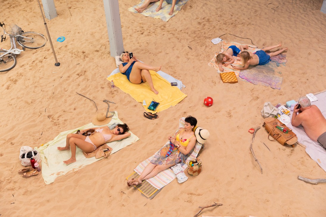 Multiple beachgoers relaxing on sand with towels, umbrellas, and personal items on a sunny day.