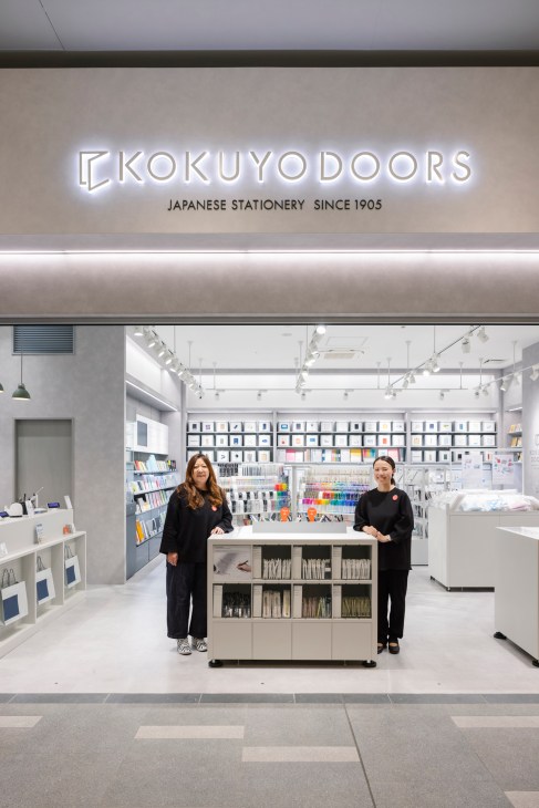 Two staff members in black uniforms stand in a bright white Kokuyo Doors stationery store with organized displays