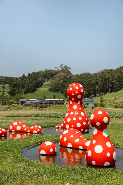 Red polka-dotted mushroom sculptures by Yayoi Kusama scattered across a water feature in rural Japanese landscape