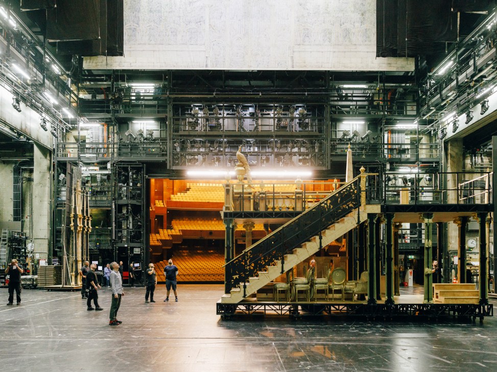 Backstage area of Deutsche Oper with industrial stage machinery, orange seating visible in background, crew preparing
