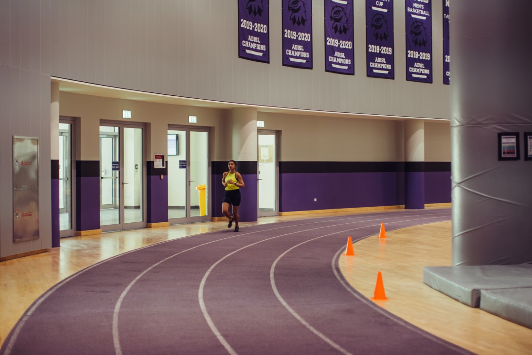 Athlete running on an indoor track with purple and cream walls and championship banners displayed above.