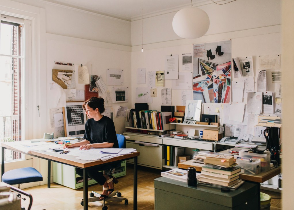 Mónica Rivera works at her desk in a bright studio surrounded by architectural sketches and design materials.