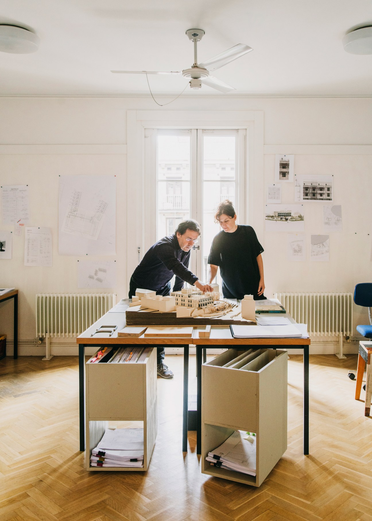 Two architects reviewing architectural models and plans at a wooden table in a bright studio office.