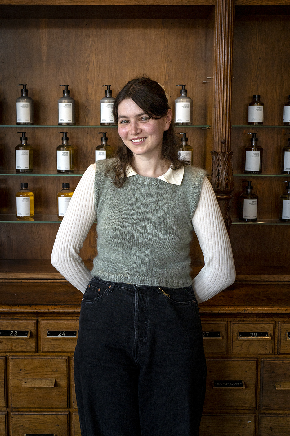 A smiling woman with brown hair wearing a gray sweater vest and white shirt standing in front of wooden shelves