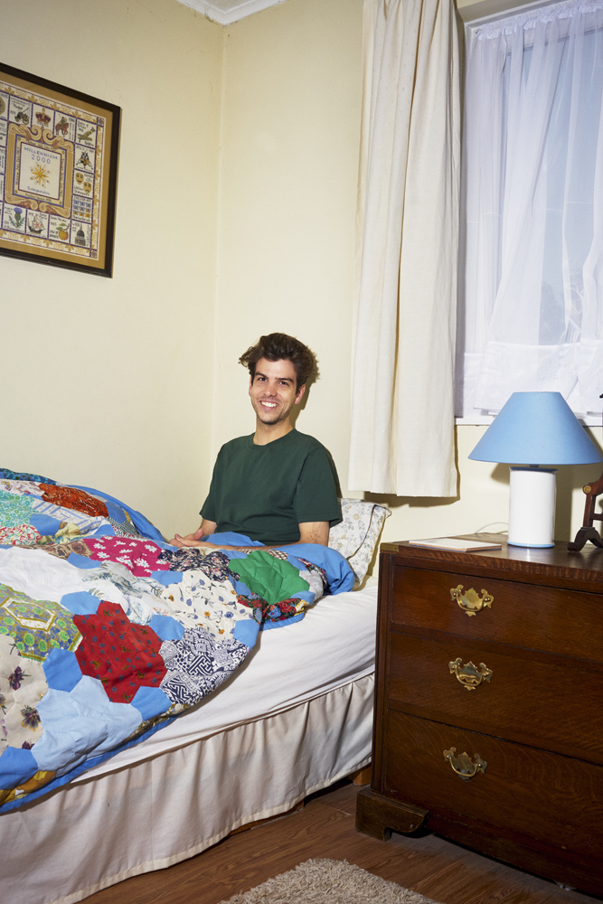 Man sitting on a bed with a colorful patchwork quilt in a modest bedroom with pale yellow walls and a blue lamp.