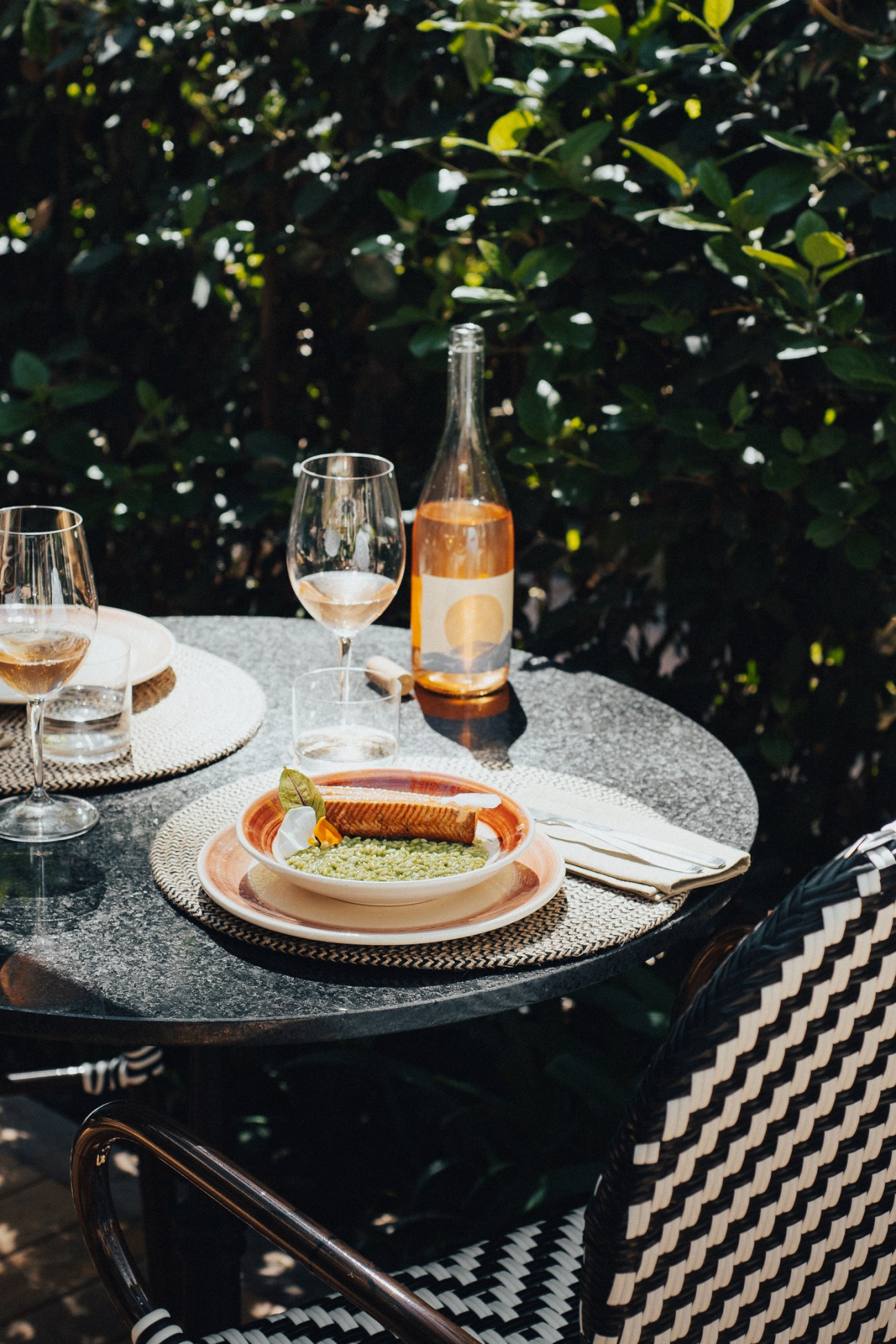 Outdoor dining table set with rosé wine, plated fish dish, and glasses under leafy green canopy.