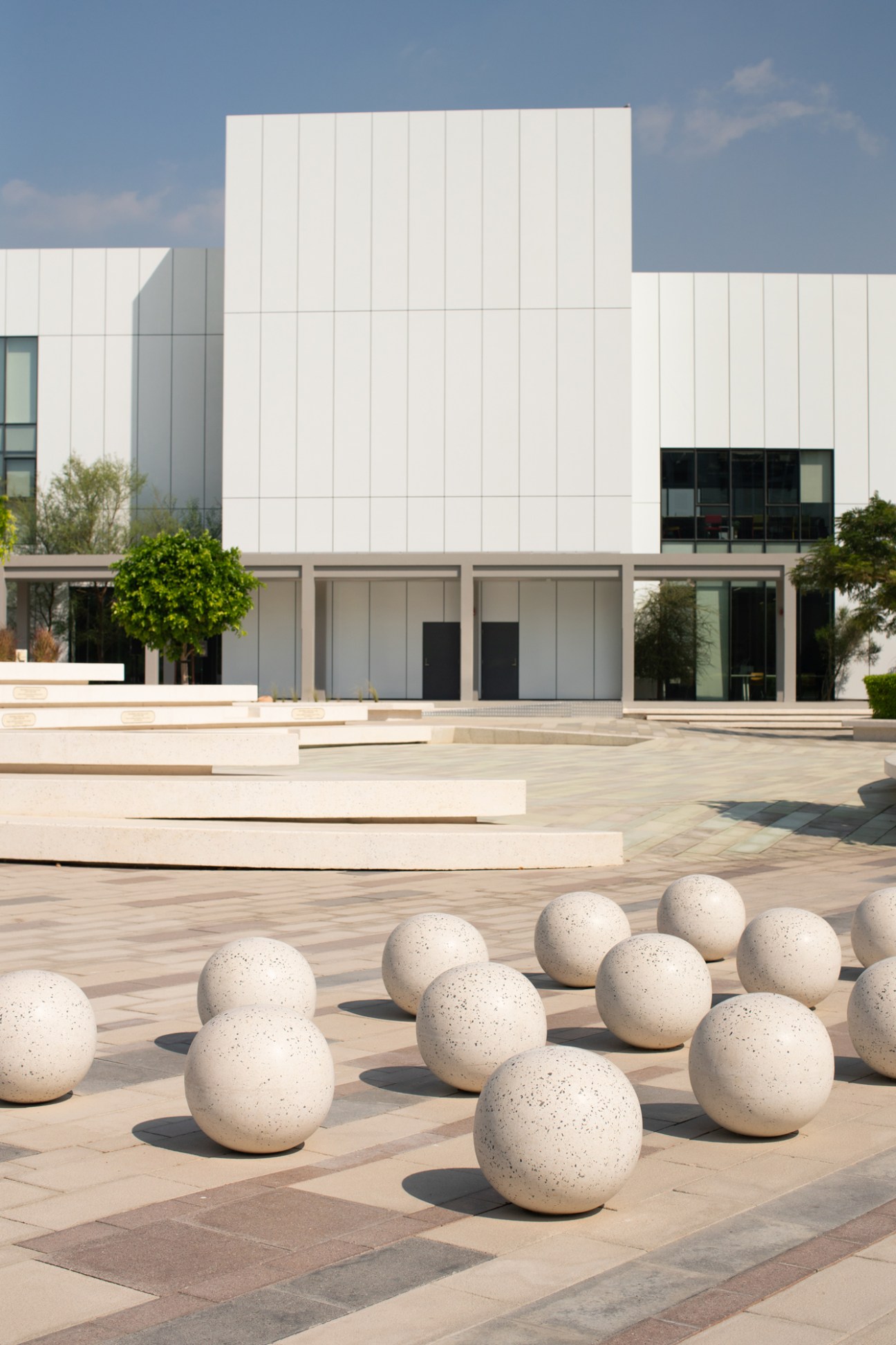 Modern white-paneled office building with ground-floor retail spaces and stone spheres in plaza.