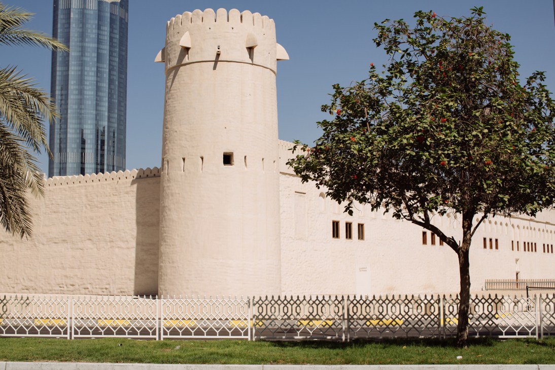 A historic white fortress with crenellated walls and tower contrasts with a modern glass building.