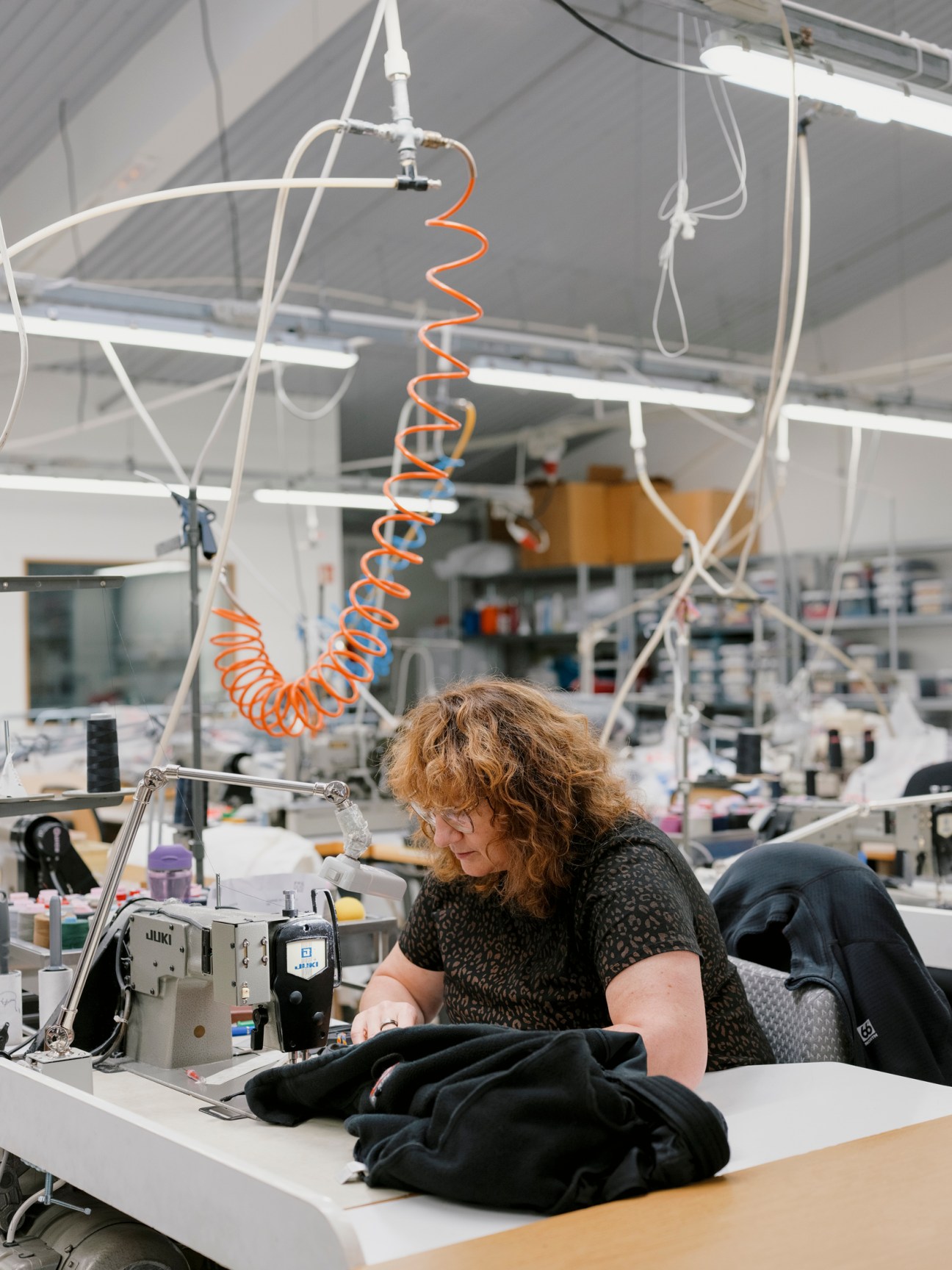 Woman with curly hair working at sewing machine repairing dark clothing in 66°North workshop