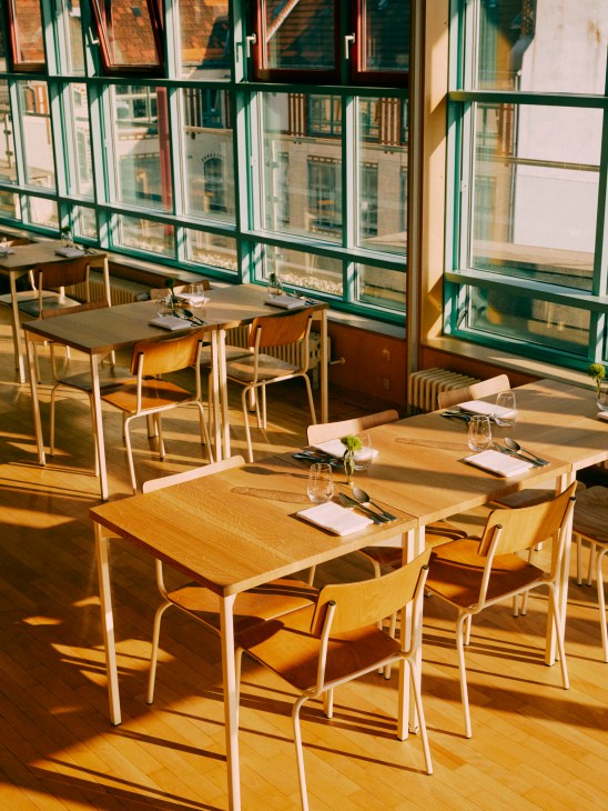 Modern restaurant dining room with wooden tables and chairs bathed in golden sunlight from large windows