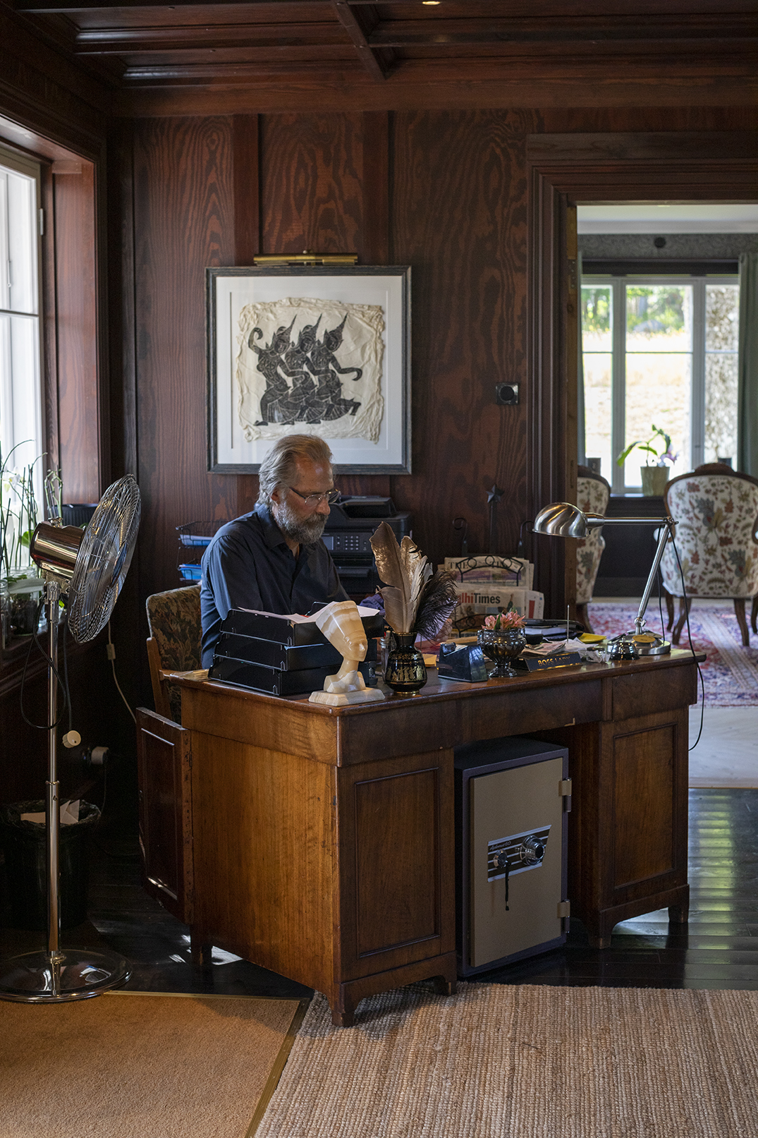 Man with beard and glasses reading at wooden desk in wood-paneled study with quill pen and vintage decor