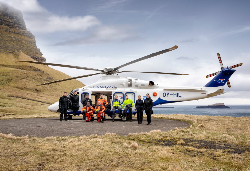 White and blue helicopter with crew members in safety gear standing beside it on grassy terrain