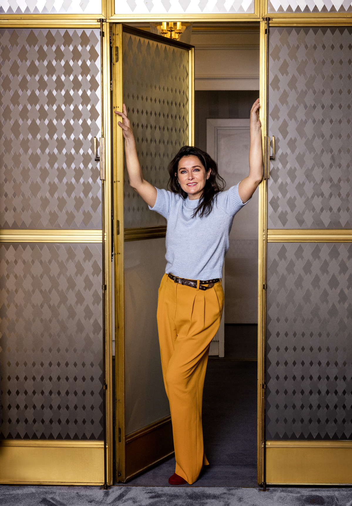 Sidse Babett Knudsen standing in doorway of ornate gold-framed glass doors with geometric patterned walls