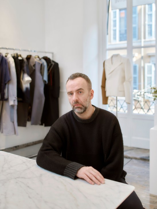 Man in dark sweater sitting at white table in minimalist fashion studio with clothing racks in background