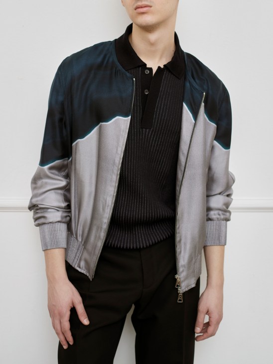 Model wearing layered outfit with color-blocked bomber jacket, black ribbed polo sweater and dark trousers