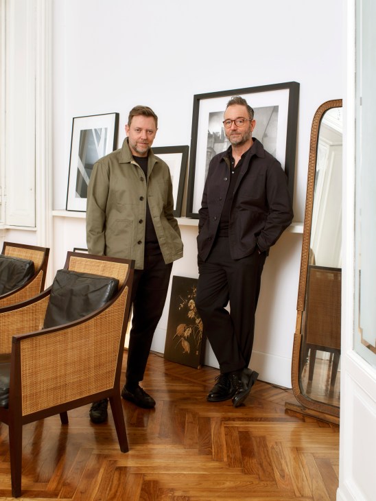 Two men in stylish casual wear stand in an elegant room with framed artwork, wooden chairs, and parquet flooring