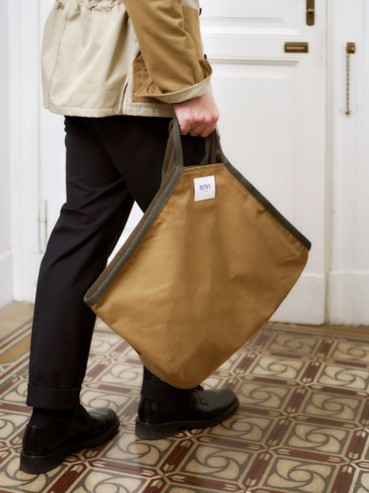 Person carrying a mustard-colored tote bag with ROVI label, wearing beige jacket and black trousers on patterned floor