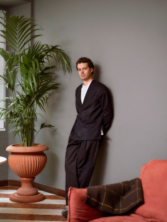 Man in black kimono-style jacket and trousers standing against grey wall next to potted palm plant and coral sofa