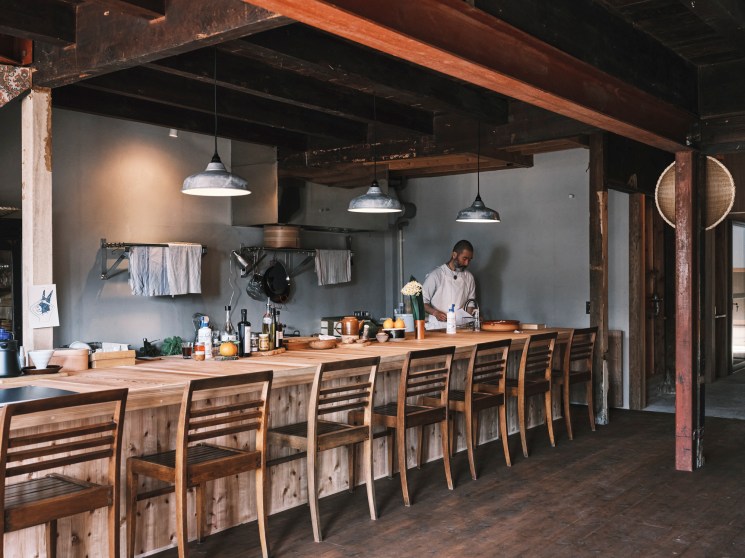 Chef preparing food behind a long wooden chef's table in a rustic kitchen.