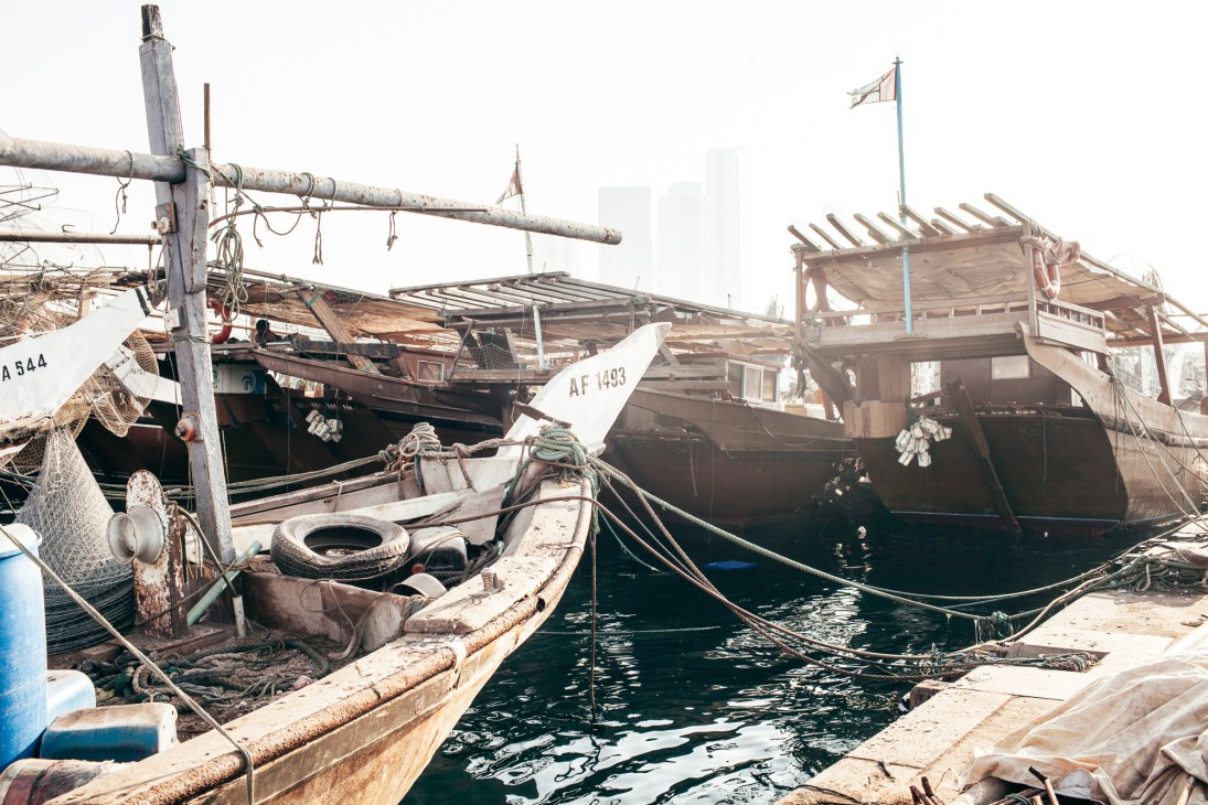 Weathered fishing boats moored at a harbor with ropes and buoys, cityscape in background.