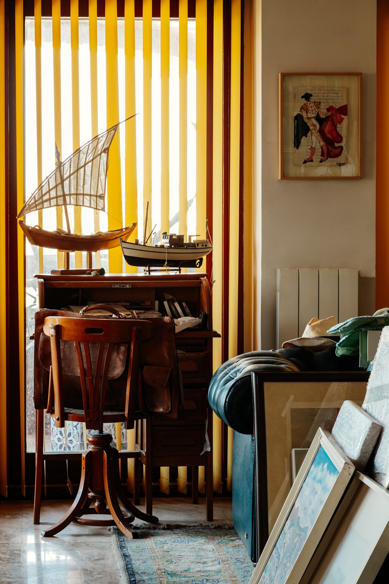 Room with a desk and chair, with ships standing on the desk, a frames picture on the wall and more framed pictures standing on the floor