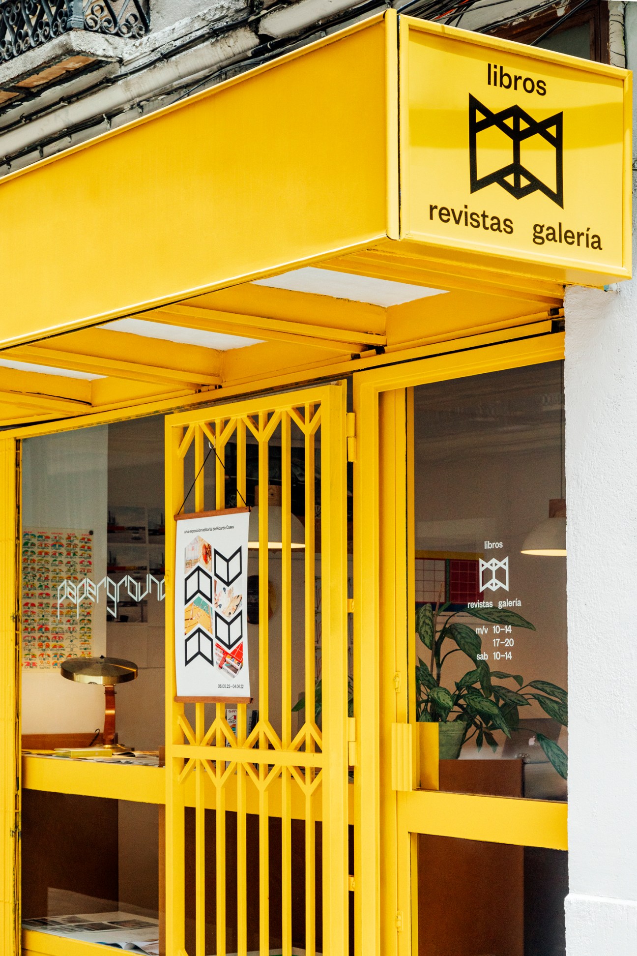 Yellow storefront of Paperground bookstore in Madrid with geometric logo and operating hours on window