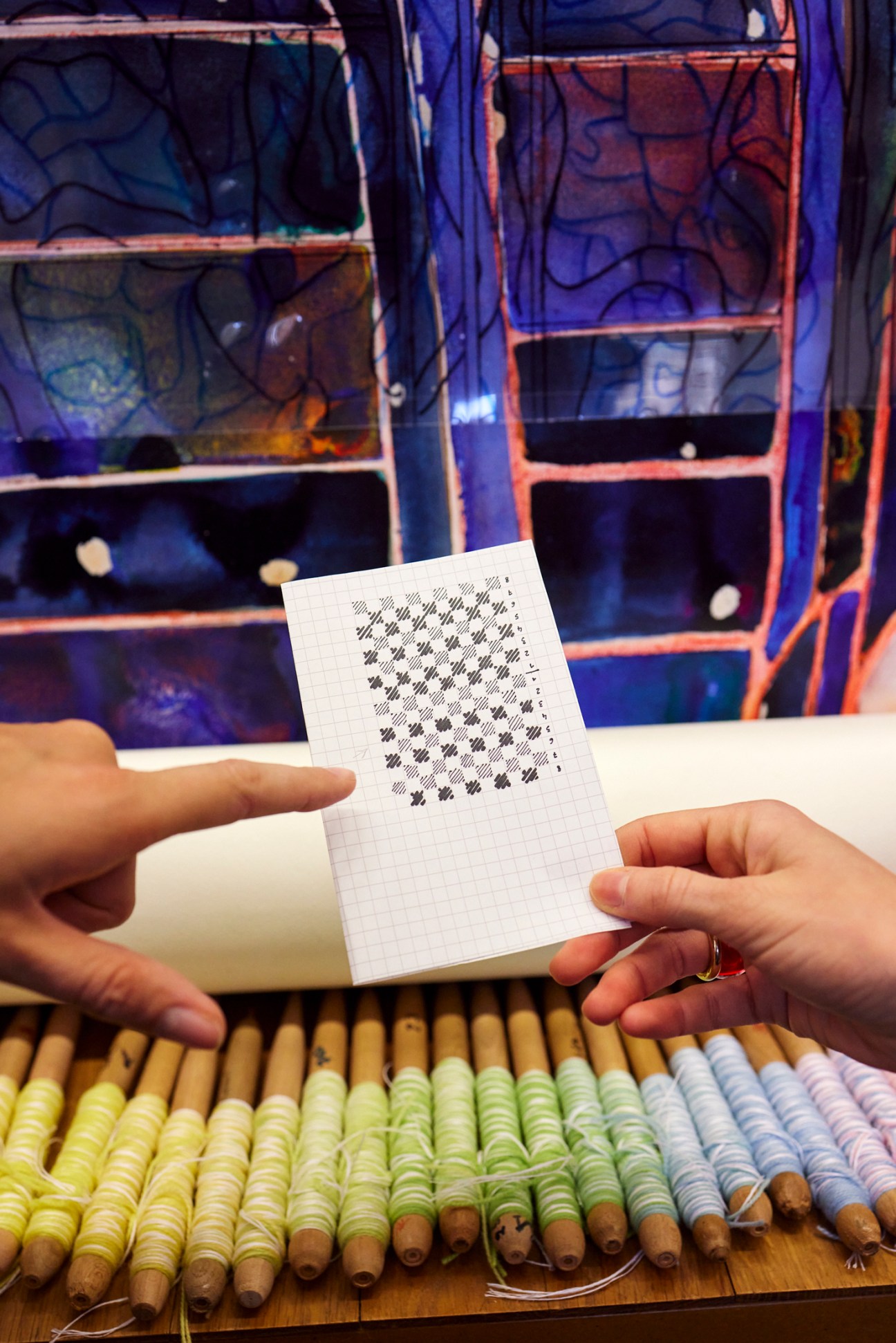 Hands holding a grid-patterned notebook with dot pattern design, displayed in front of colorful pencils and blue shelving.