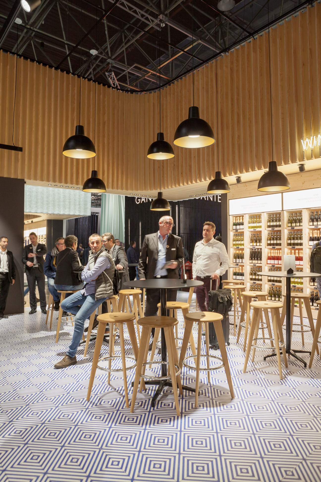Interior of The Village food market at EuroShop featuring pendant lights, wooden walls, bar seating, and visitors browsing wine shelves.
