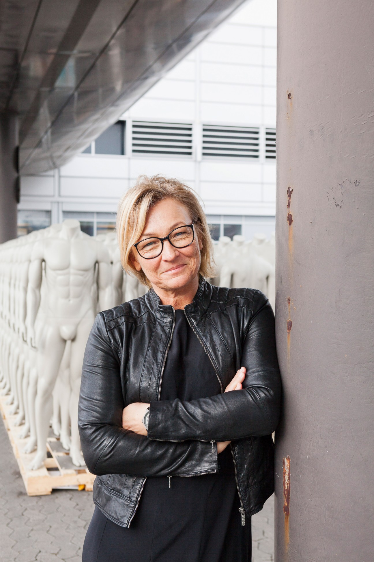Woman with blonde hair wearing glasses and a black leather jacket stands against a concrete wall.