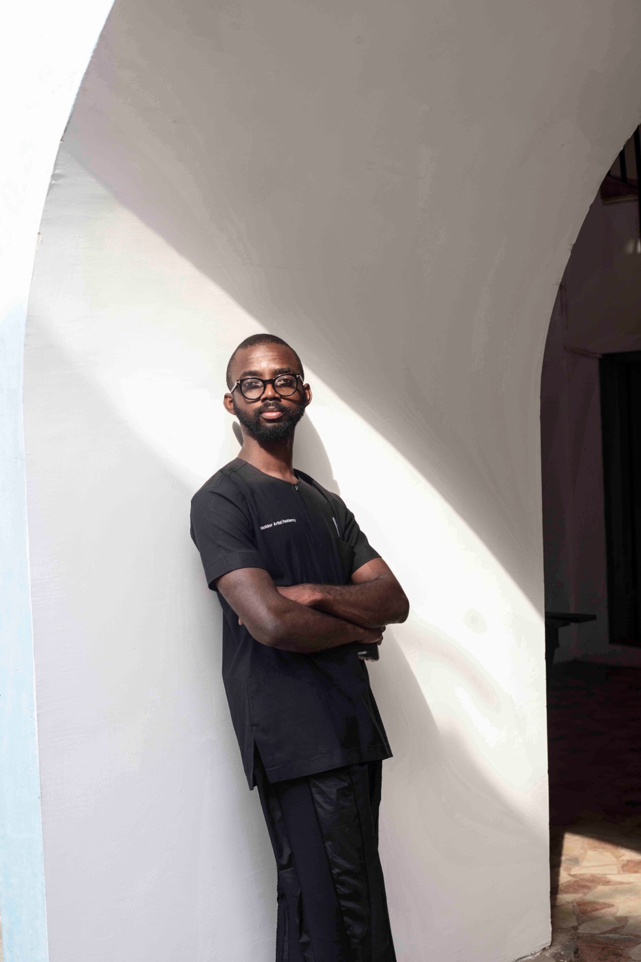 Joseph Awuah-Darko standing with arms crossed in black clothing against white wall