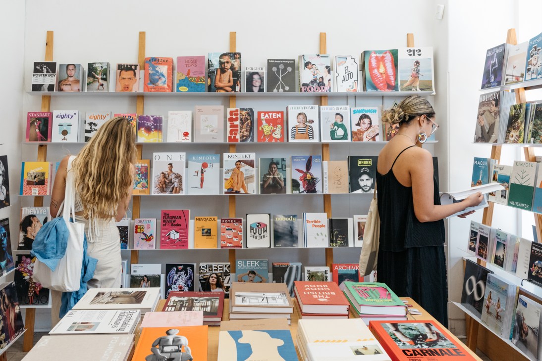Two women browsing magazines and books on wooden shelves displaying various publications