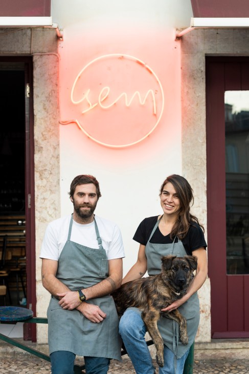 Two chefs in aprons sitting outside Sem Restaurant in Lisbon with their dog under a neon sign