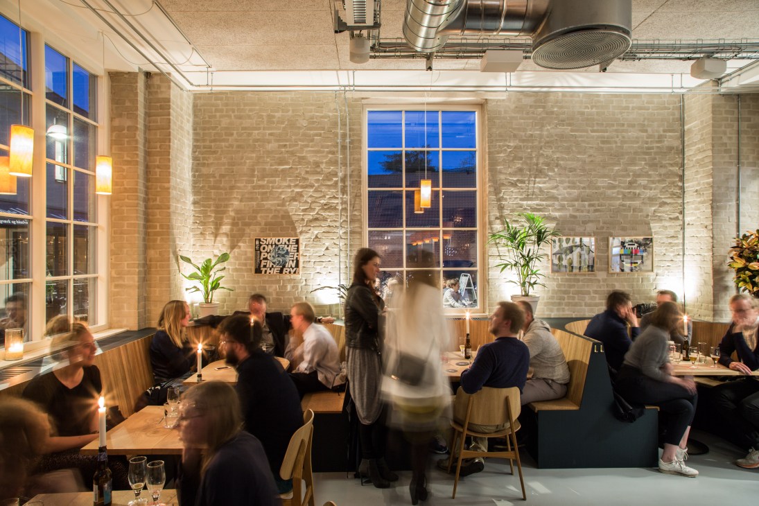 Interior of Brus pub in Copenhagen with diners at tables, exposed brick walls, large windows, and pendant lighting.