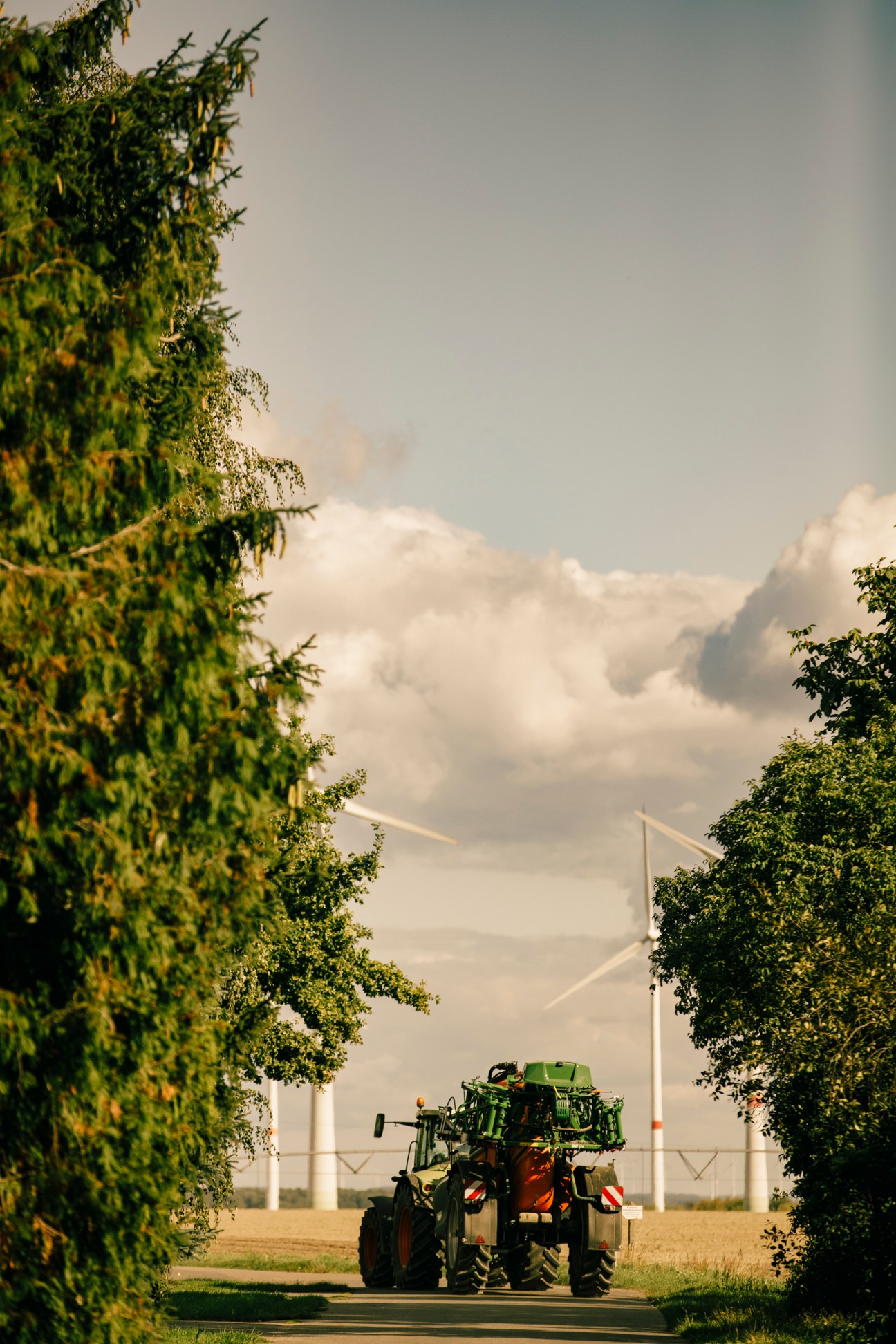 Green tractor driving down rural road lined with trees and wind turbines visible in background