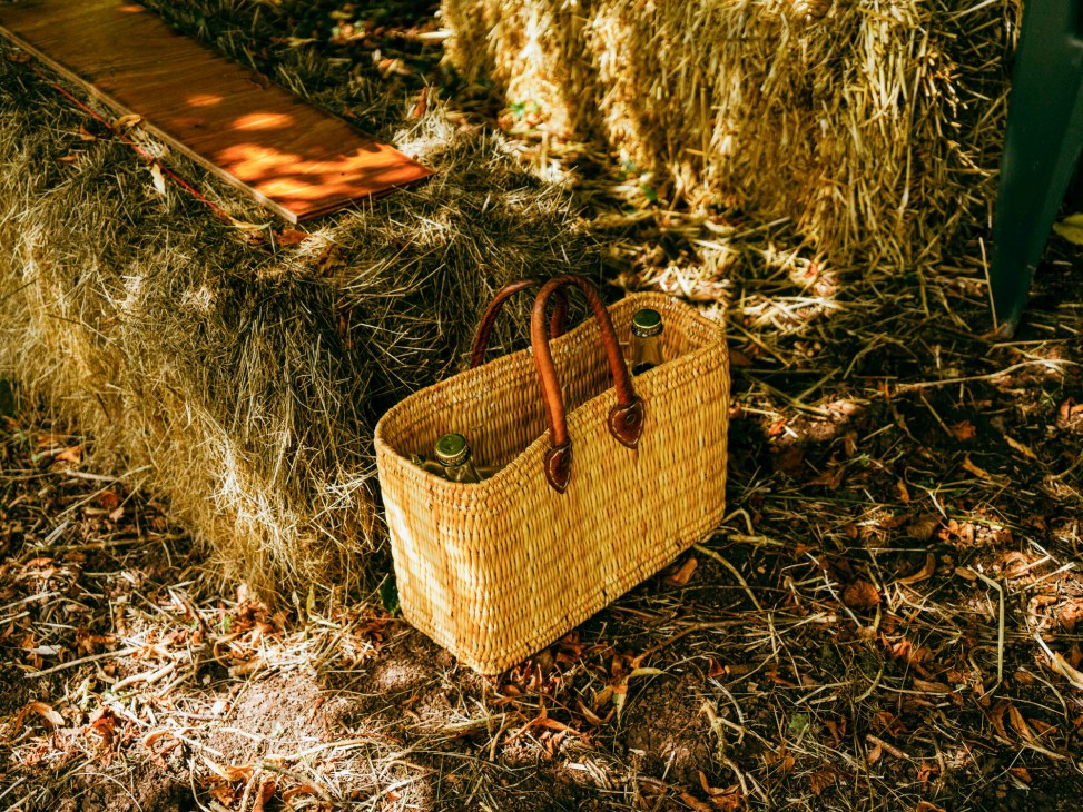 Woven basket with leather handles containing two bottles sitting on straw-covered ground near hay bales