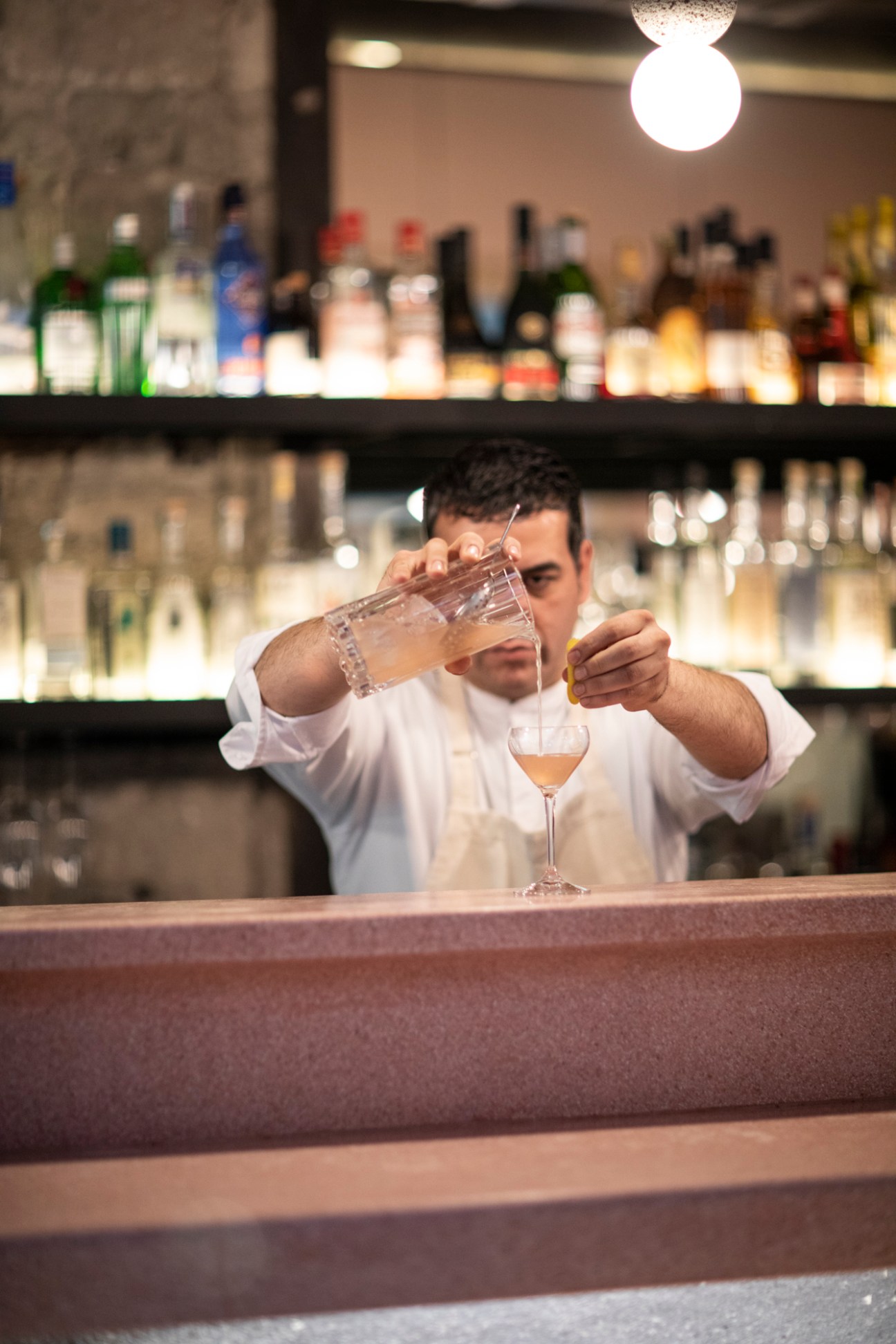 Bartender in white shirt pouring a cocktail from mixing glass into stemmed glass behind bar