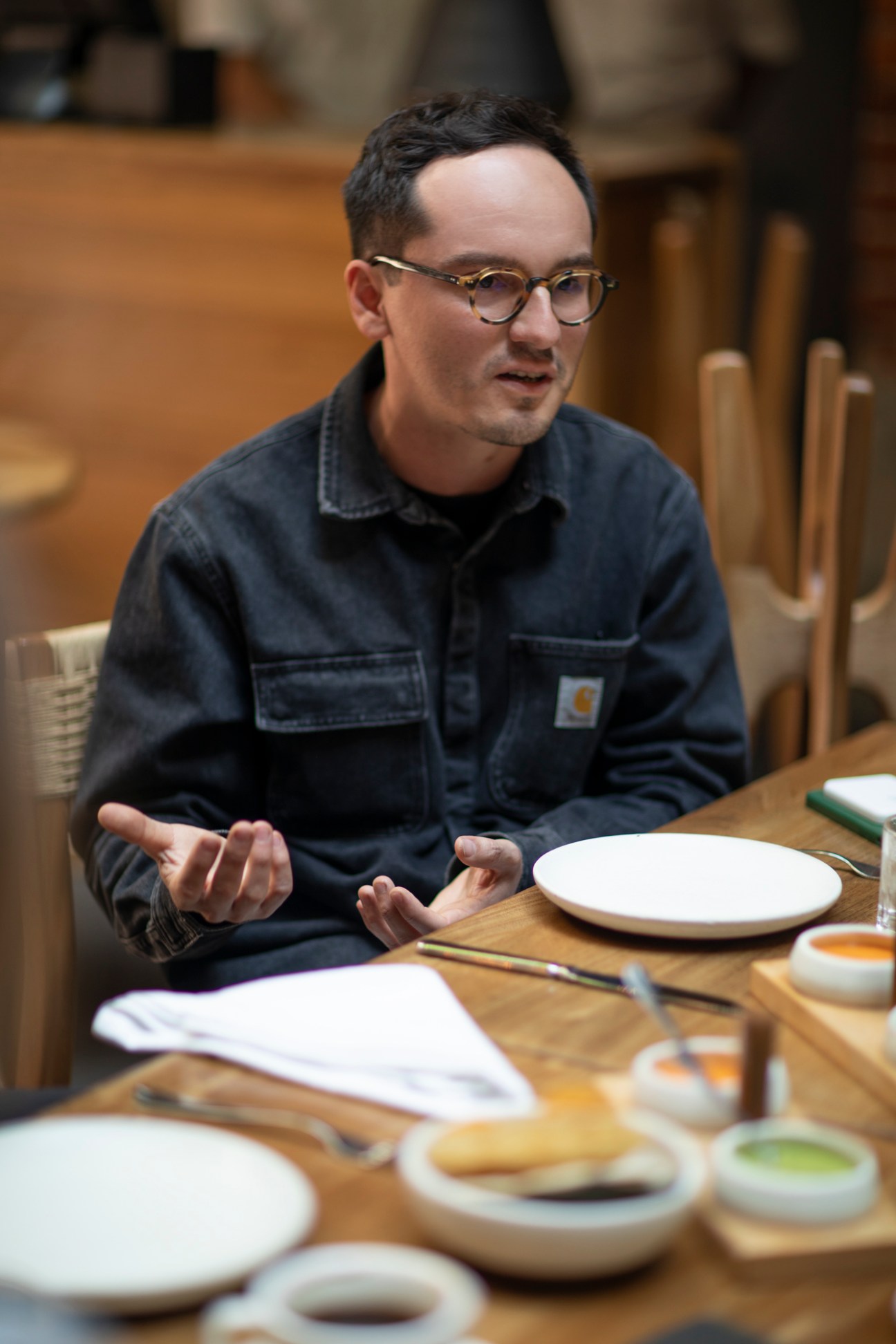 Man in dark collared shirt and round glasses gesturing while speaking at restaurant table with plates